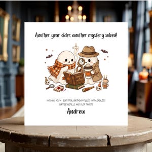 Könnte beinhalten: Eine quadratische Grußkarte mit zwei Cartoon-Geistern. Einer trägt einen karierten Schal, der andere einen Fedora und Trenchcoat mit Lupe und Kaffeebecher. Text: Another year older, another mystery solved! Wishing you a boo-tiful birthday filled with endless coffee refills and plot twists. Andrew.