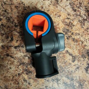 Può includere: Un morsetto di fissaggio in plastica nera con un'apertura circolare, un inserto quadrato arancione brillante e un anello blu. Il dispositivo include una manopola di regolazione grigia laterale e una base cilindrica con un dettaglio a coste.