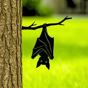 May include: A black bat silhouette hangs upside down from a tree branch against a bright green lawn. The bat's wings are spread, and its ears are pointed. The tree trunk is brown and textured. The word "LUXLAYERED" is at the bottom right.