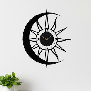 May include: Black metal wall clock featuring a sun and crescent moon design. The clock face has gold-colored hands and numerals. The clock is mounted on a white wall. The text "LUXLAYERED" is at the bottom.