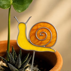 May include: A stained-glass snail garden stake with a yellow body and an orange shell, outlined in silver. The stake is in a terracotta pot with a succulent plant. The snail design is a decorative garden ornament.