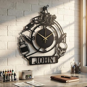 May include: A black metal wall clock featuring a tattoo machine, ink bottle, and skull design. The clock face has gold hands and numbers 3, 6, 9, and 12. A rectangular nameplate at the bottom displays the name JOHN in bold, cut-out capital letters.