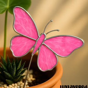 Op de afbeelding: Een roze glas-in-lood vlinder tuinpaal met een zilverkleurig metalen frame. De vlinder staat in een terracotta pot met een vetplant. De tekst "LUXLAYEREDred" is zichtbaar.