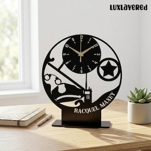 May include: Black laser-cut desk clock with a police theme. The clock features a circular design with a star, handcuffs, a radio, and the name "RACQUEL MAXEY". The clock hands are gold-colored, and the text "LuxLayered" is at the top.
