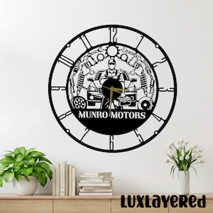 May include: Black metal wall clock with a car mechanic design. The clock features a car, tools, and the text "MUNRO MOTORS". Roman numerals mark the hours, and the clock hands are gold.