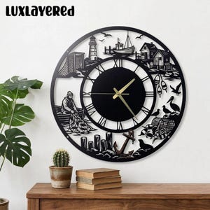 Könnte beinhalten: Schwarze Metall-Wanduhr mit nautischem Thema. Die Uhr zeigt einen Leuchtturm, ein Boot, einen Fischer und andere Küstenelemente. Die Zeiger der Uhr sind goldfarben. Der Text "LUXLAYERED" befindet sich oben auf der Uhr.