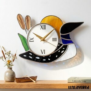 Puede incluir: Un reloj de pared de vidrieras con un diseño de pato, con una esfera blanca y manecillas doradas. El reloj tiene un sol amarillo, un cuerpo negro y blanco y detalles azules. El reloj tiene aproximadamente 30 cm de diámetro.