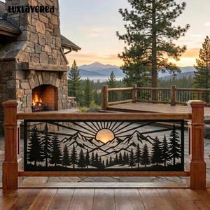 May include: A black metal railing panel featuring a laser-cut design of a mountain range, a sunburst, and a forest of pine trees. The panel is installed between two wooden posts on a deck, with a stone fireplace and mountain view behind it.