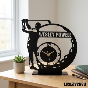 May include: Black metal golf-themed clock with a golfer silhouette, a golf ball design, and the name "WESLEY POWELL". The clock face has gold hands and a simple numeral layout. The clock is on a wooden surface.