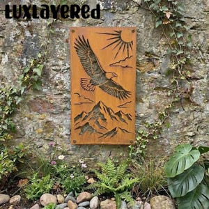 May include: A rectangular, rusty-brown metal wall art piece featuring a soaring eagle, sun, clouds, and mountain range cut-out design. The artwork is mounted on a stone wall, surrounded by greenery and the text "luxlayeredred" is at the top.