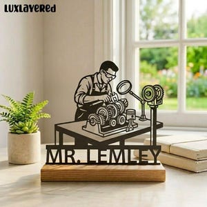 Może przedstawiać: Czarna metalowa rzeźba przedstawiająca mechanika pracującego przy silniku, zamontowana na drewnianej podstawie. Na froncie widnieje napis "MR. LEMLEY". Tło przedstawia okno i małą roślinę doniczkową.