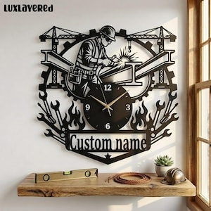 May include: A black metal wall clock featuring a silhouette of a construction worker welding steel beams, surrounded by cranes, gears, and wrenches. The clock face has gold hands and numbers, with a banner at the bottom displaying the text Custom name.