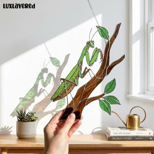 May include: A stained glass suncatcher depicting a green praying mantis perched on a brown branch with green leaves. The piece is constructed with soldered metal lines and hangs from a thin chain, casting a colorful shadow on the wall behind it.