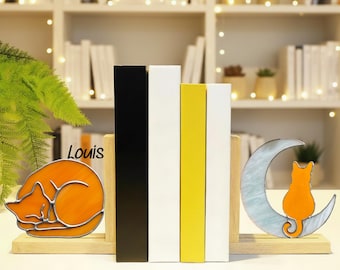 Puede incluir: Juego de sujetalibros decorativos con un gato naranja durmiendo y una silueta de gato en una luna creciente. Los sujetalibros se combinan con libros negros, blancos y amarillos. La palabra "Louis" es visible.