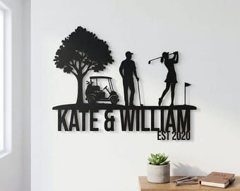 May include: Black metal golf-themed wall art featuring a tree, golf cart, and two golfers. The names "KATE & WILLIAM" and the year "EST 2020" are displayed below the figures. A decorative piece for a golf enthusiast.