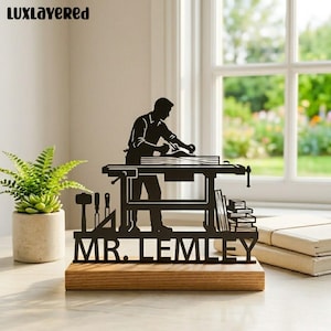 Könnte beinhalten: Schwarze Metall-Silhouette eines Zimmermanns auf einem Holzsockel. Das Design zeigt einen Zimmermann, der an einer Werkbank arbeitet, mit Werkzeugen und dem Text "MR. LEMLEY". Links ist eine kleine Pflanze im Topf.