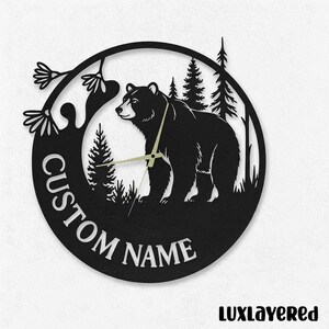 May include: Black metal wall clock featuring a bear silhouette in a forest scene. The clock has a circular design with the words "CUSTOM NAME" and a clock face with hands. The clock is a decorative item.