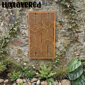 May include: A rectangular, rust-colored metal wall art panel with a repeating arched design. The panel is mounted on a weathered stone wall, surrounded by greenery and foliage. The text "luxlayered" is at the top.