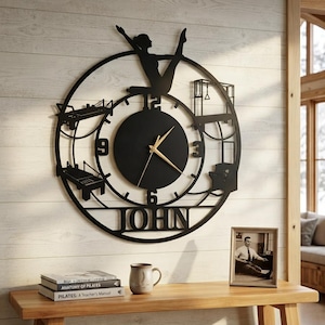 May include: A circular black metal wall clock featuring a silhouette of a person performing a pilates pose at the top. The clock face includes gold hands, numbers 3, 6, 9, and 12, and cutouts of pilates equipment. The name JOHN is cut out at the bottom.