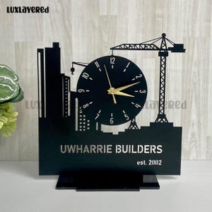 May include: Black desk clock featuring a cityscape silhouette with a clock face and construction crane. The clock has gold hands and the text "Uwharrie Builders est. 2002". The clock is approximately 8 inches tall.