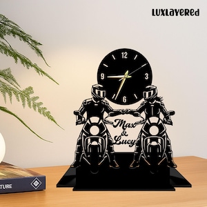 Könnte beinhalten: Schwarze Uhr zum Thema Motorrad mit zwei Fahrern und einem kreisförmigen Zifferblatt. Die Uhr trägt die Namen "Max & Lucy" und den Markennamen "LUXLAYERED". Sie steht auf einem schwarzen Sockel und liegt auf einer Holzoberfläche.