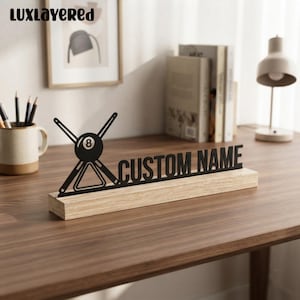 May include: A black metal desk sign featuring a billiard 8-ball graphic with crossed cues and a triangle rack, mounted on a rectangular light wood base. The sign displays the text CUSTOM NAME in bold, uppercase, sans-serif black lettering.