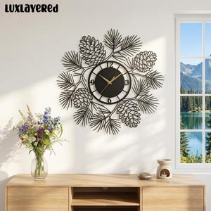 May include: A black metal wall clock featuring a circular face with gold hands and numbers 3, 6, 9, and 12. The clock is surrounded by a decorative frame of laser-cut pine needles and pinecones. The text LUXLAYERED is visible in the top left corner.