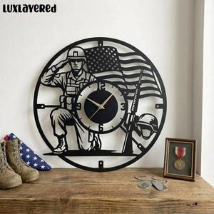 Puede incluir: Reloj de pared de metal negro con un soldado saludando, la bandera estadounidense, un rifle y un casco. Las manecillas del reloj son negras y la esfera tiene los números 12, 3, 6 y 9. El reloj está sobre una superficie de madera.