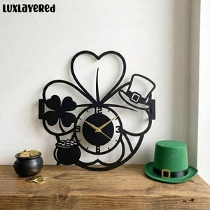 May include: Black metal wall clock in the shape of a four-leaf clover, with a clock face, a leprechaun hat, and a pot of gold. A green leprechaun hat sits on a wooden surface. The clock hands are gold. The brand name "LUXLayered" is at the top.