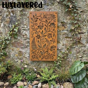 Puede incluir: Un panel de jardín rectangular de metal color óxido con un diseño floral. El panel está montado en una pared de piedra y rodeado de vegetación. El texto "luxlayered" está en la parte superior.