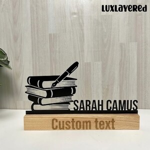 Może przedstawiać: Czarny organizer biurkowy z stosu książek i piórem, z imieniem "SARAH CAMUS" i słowami "Custom text" wygrawerowanymi na drewnianej podstawie. Logo "LuxLayered" znajduje się w prawym górnym rogu.