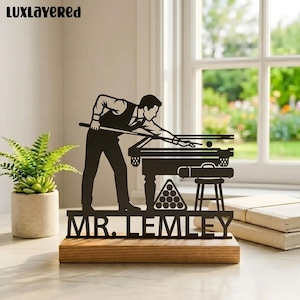 May include: A black metal billiards-themed decorative piece. It features a silhouette of a person playing pool, a pool table, and the text "MR. LEMLEY" on a wooden base. A small plant in a pot is on the left.