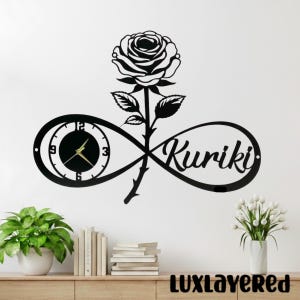 May include: Black metal wall clock featuring a rose and infinity symbol design. The clock face is round with gold hands and numerals. The word "Kuriki" is written in cursive. The clock is mounted on a white wall.