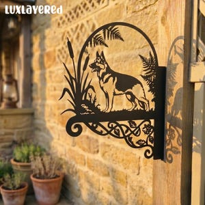 Op de afbeelding: Zwarte metalen wandkunst met een silhouet van een Duitse herdershond in een boogvormig frame. Het ontwerp omvat lisdodden, varens en decoratieve krullen. De tekst "LUXLAYERED" is bovenaan zichtbaar.