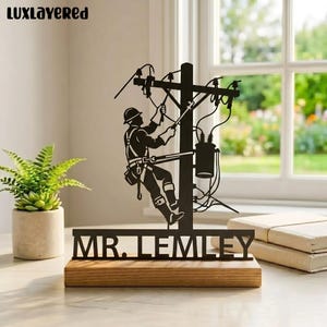 May include: Black metal silhouette of a lineman working on a power pole, mounted on a wooden base. The name "MR. LEMLEY" is cut out below the lineman. The image is set against a window with a view of greenery.