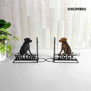May include: Black metal bookends featuring silhouettes of a black and a brown Labrador Retriever. The bookends have the names "WILLOW" and "ZIGGY" cut out at the base. The background is a light wood grain pattern.