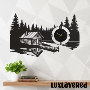 Puede incluir: Reloj de pared de metal negro con una escena de cabaña con un lago, árboles y un muelle. La esfera del reloj es negra con manecillas doradas y números blancos. El reloj está montado en una pared blanca.