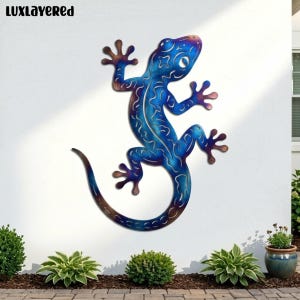 May include: A blue metal lizard wall art piece with a gradient of colors. The lizard is mounted on a white wall. The lizard's body has intricate cut-out designs. The text "LUXLAYERED" is in the top left corner.