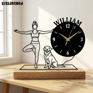 Può includere: Un orologio da tavolo in metallo nero con la sagoma di una persona in posizione yoga dell'albero e un cane seduto su un tappetino. Il quadrante è circolare con il nome WILLIAM stampato in alto, lancette dorate e una base in legno rettangolare.