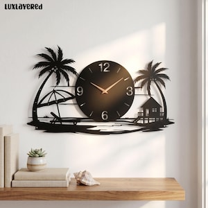 May include: A black metal wall clock featuring a tropical beach scene with two palm trees, a beach umbrella, a lounge chair, and a small hut. The circular clock face has gold-colored hands and the numbers 3, 6, 9, and 12 in a metallic finish.