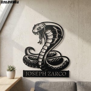 Puede incluir: Arte mural de metal negro de una cobra en una pose llamativa, con su capucha extendida y colmillos a la vista. El nombre "JOSEPH ZARCO" se muestra en una placa rectangular debajo de la serpiente. La obra está montada en una pared clara.
