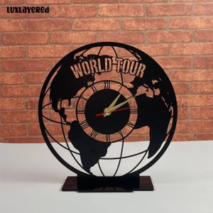 May include: Black World Tour clock with a globe design. The clock features Roman numerals and gold-colored hands. The clock is mounted on a dark brown rectangular base. The text "WORLD TOUR" is displayed at the top of the clock.