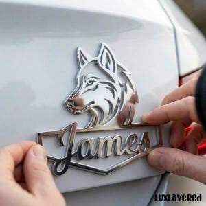 Könnte beinhalten: Ein silbernes Autoemblem mit einem Wolfskopf über dem Namen "James" in einer stilisierten Schriftart. Das Emblem wird auf einer weißen Autooberfläche platziert, wobei die Hände einer Person sichtbar sind.