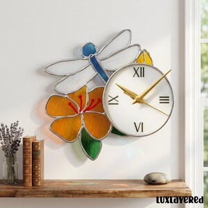 Könnte beinhalten: Eine dekorative Wanduhr mit einem Buntglasdesign einer Libelle und einer Blume. Die Uhr hat ein weißes Zifferblatt mit goldenen Zeigern und römischen Ziffern. Die Libelle ist blau und weiß, und die Blume ist orange und gelb.