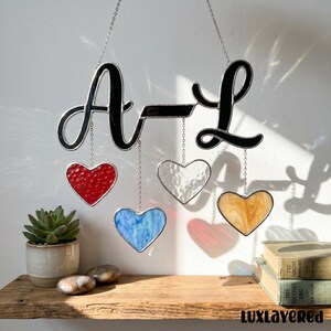 以下が含まれることがあります： 黒い筆記体で「A-L」の文字が書かれたステンドグラスの装飾品。黒いバーで繋がれています。赤、透明、青、黄色のハート型のガラスがチェーンで吊り下げられています。多肉植物と本が木製の棚に置かれています。