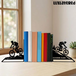 May include: Black metal bookends featuring a cyclist on a mountain bike, with a mountain silhouette design. The bookends are on a wooden surface, holding a row of colorful books. The text "CUSTOM NAME" is visible on one bookend.