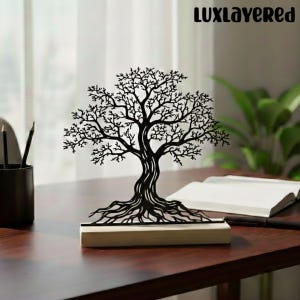 Puede incluir: Escultura de árbol de metal negro con intrincadas ramas y raíces, montada sobre una base de madera clara. La escultura se exhibe sobre un escritorio de madera oscura, con un libro abierto y lápices parcialmente visibles en el fondo. El texto "LUXLayered" es visible.