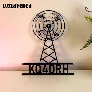 Peut inclure: Décoration murale en métal noir représentant une tour radio avec une antenne circulaire et l'indicatif d'appel KQ4ORH. Le design a une esthétique moderne et géométrique, adapté à la décoration de la maison ou du bureau. La marque "luxlayered" est visible en haut.
