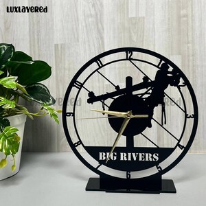 May include: Black metal clock with a circular face featuring Roman numerals and a silhouette of a helicopter. The clock hands are gold. The words "BIG RIVERS" are at the bottom of the clock face.