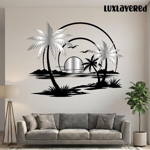 Pu&ograve; includere: Un'opera d'arte da parete in metallo nero e argento raffigurante una scena di spiaggia tropicale. Il design presenta palme, un sole al tramonto, uccelli e il testo "LUXLAYERED" nell'angolo in alto a destra. L'opera &egrave; montata su una parete di colore neutro sopra un divano grigio.